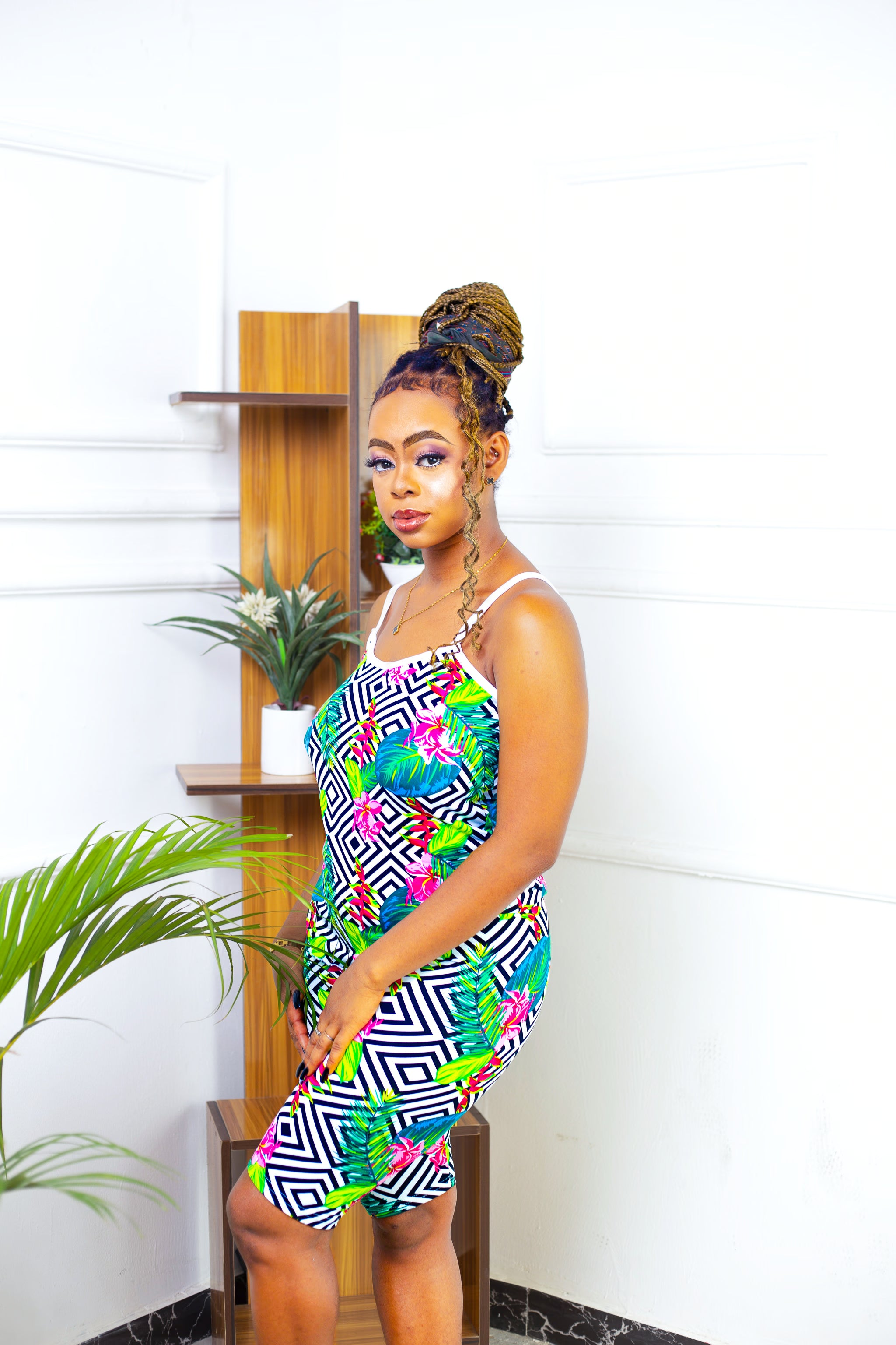 Woman wearing a colorful geometric pajama set standing indoors with a plant and wooden shelf in the background.