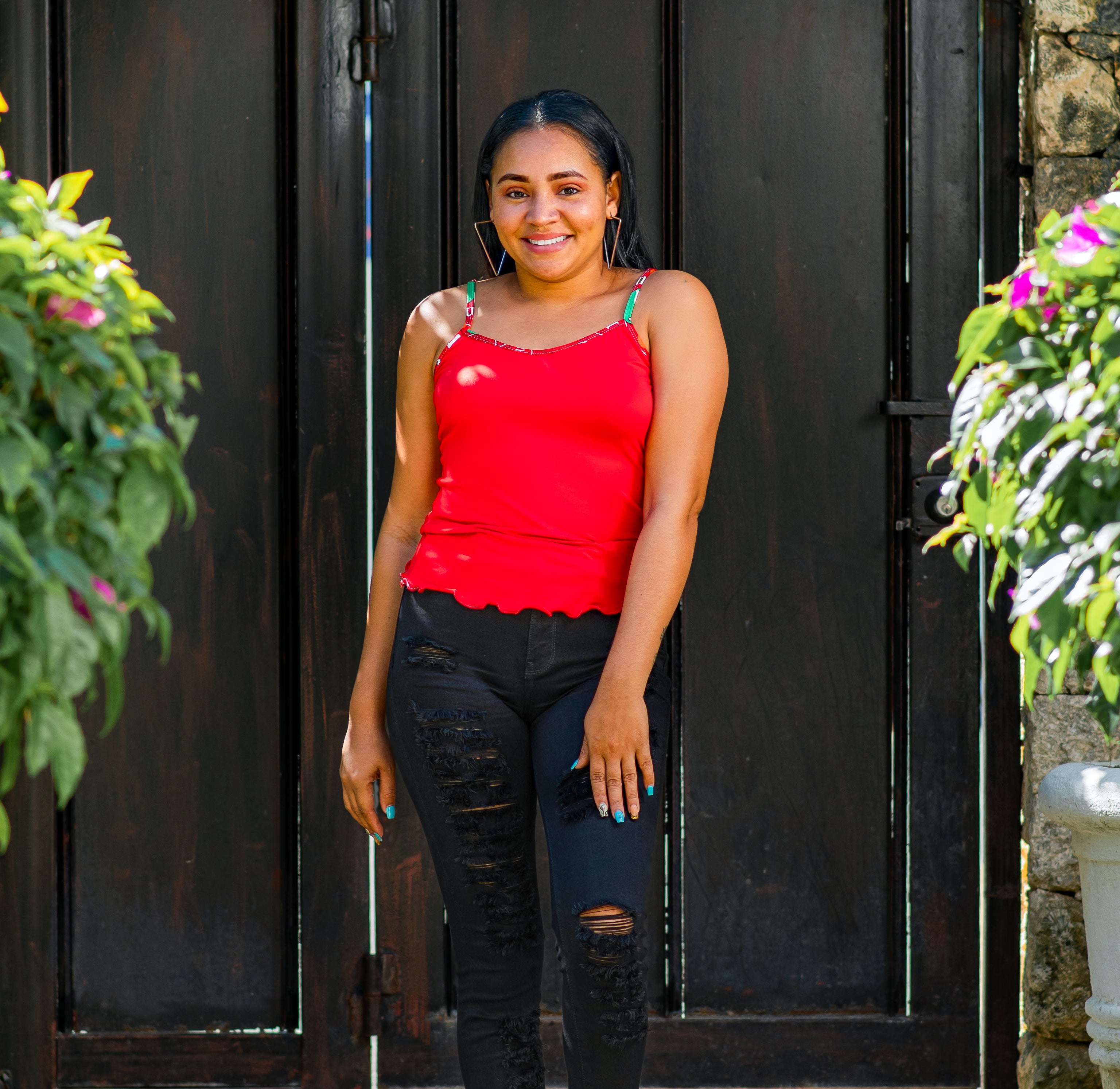 Woman in a red Cami pajama top and black jeans standing in front of a dark wooden door with plants on either side.