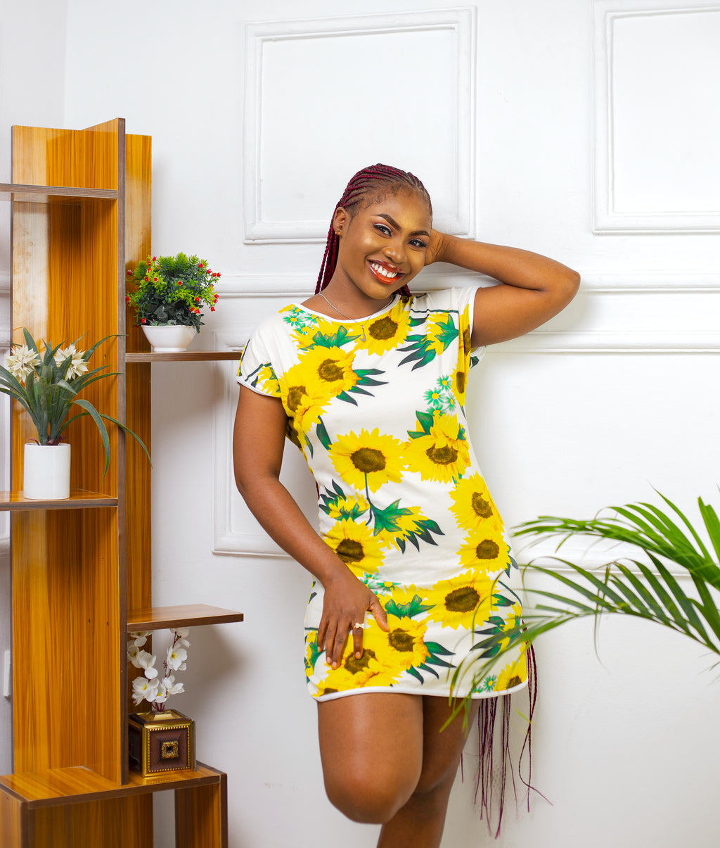 Woman wearing a Sunflower print nightgown standing in a room with white walls, plants and wooden shelves.