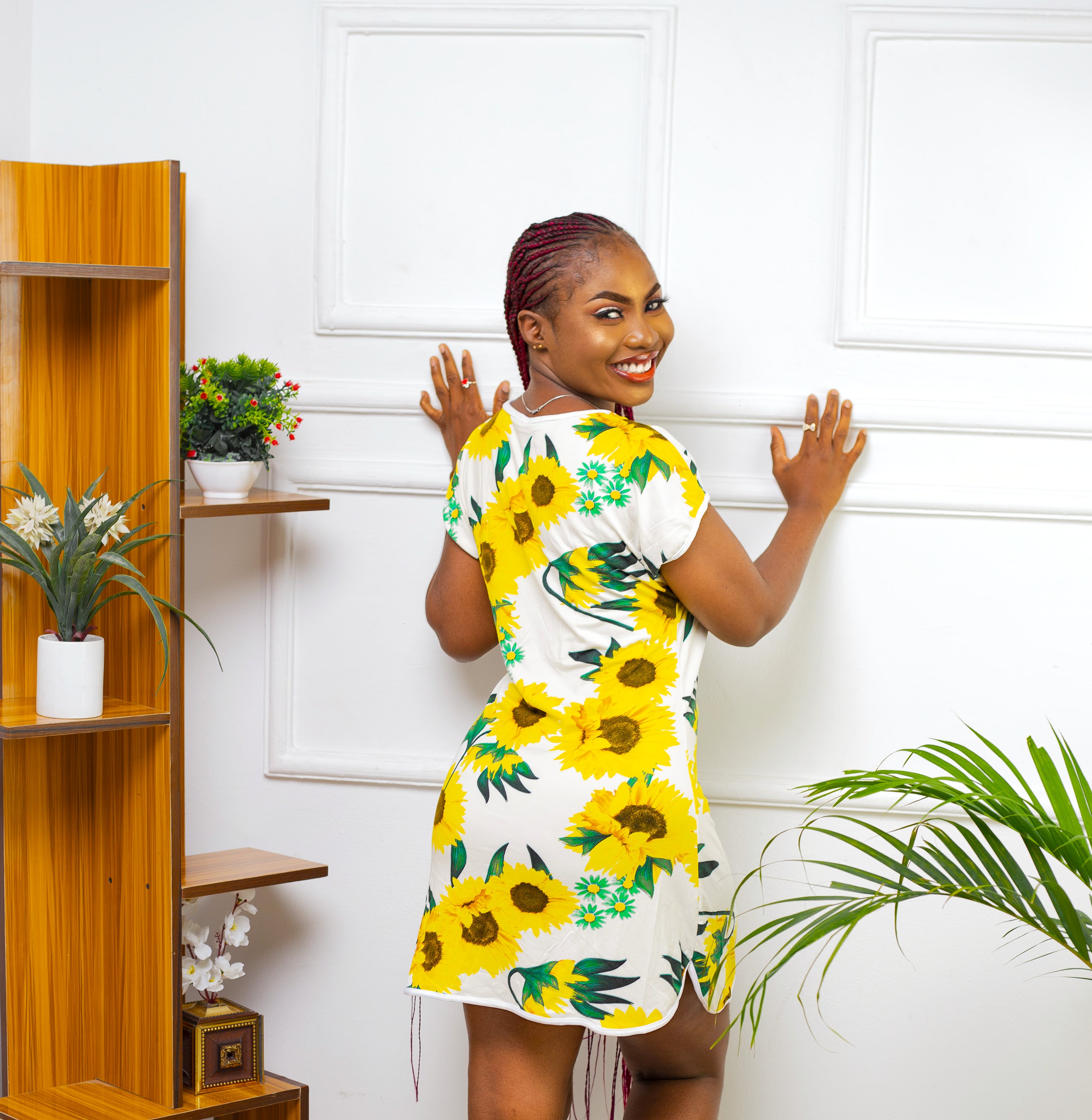 Woman wearing white sleep shirt with yellow sunflower print featuring side splits  a room with plants and shelves.