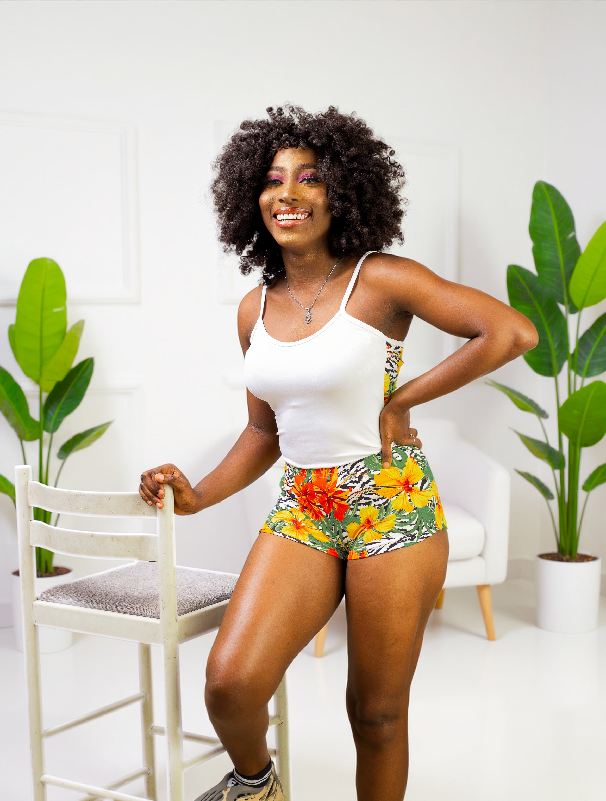 Woman in a white in the front and hibiscus flower print on back cami top and matching floral shorts standing in a room with plants.