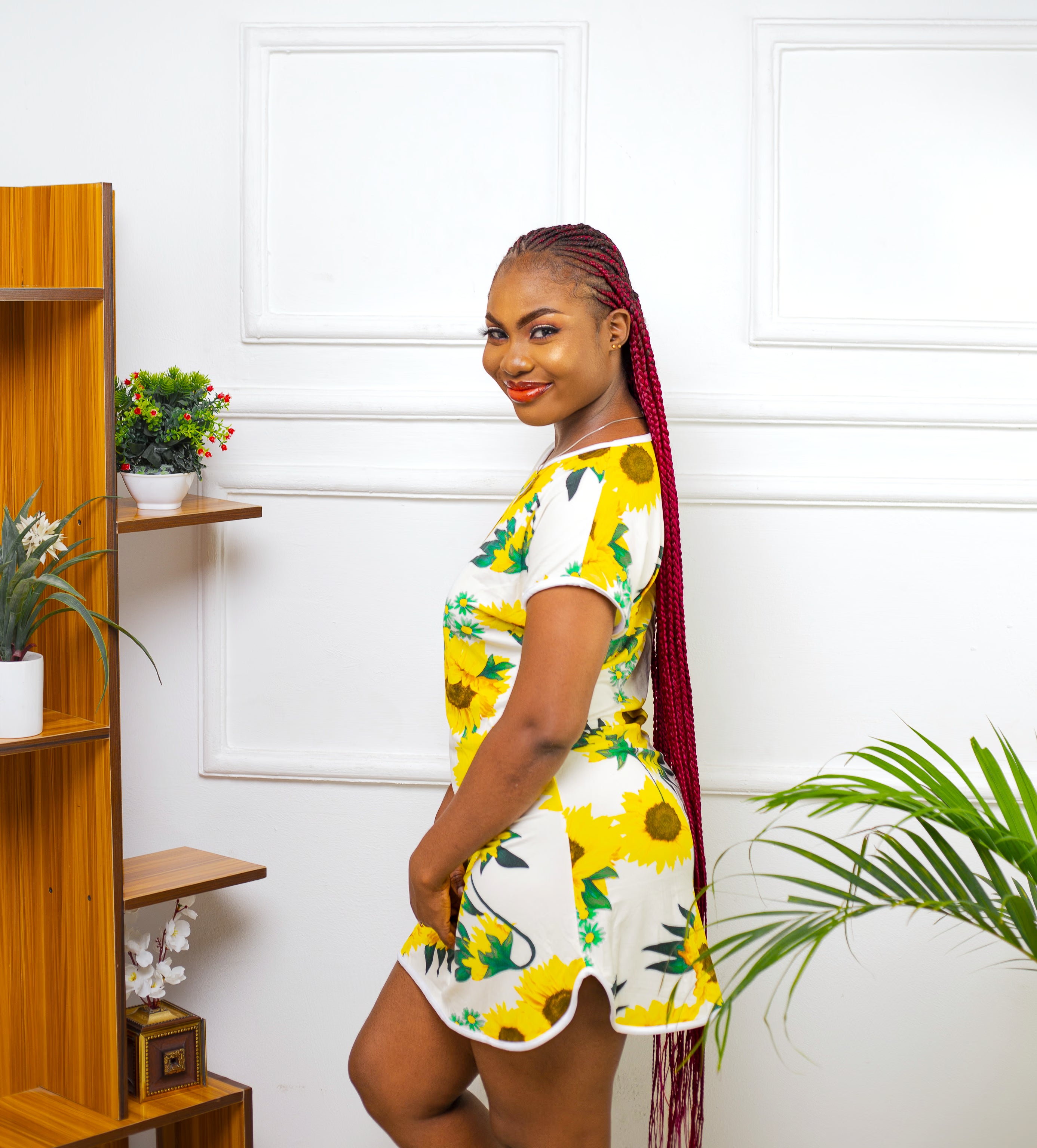 Woman wearing cheerful sunflower sleepwear dress with short sleeves, side splits, and performance fabric standing in a room with a wooden shelf and plants.